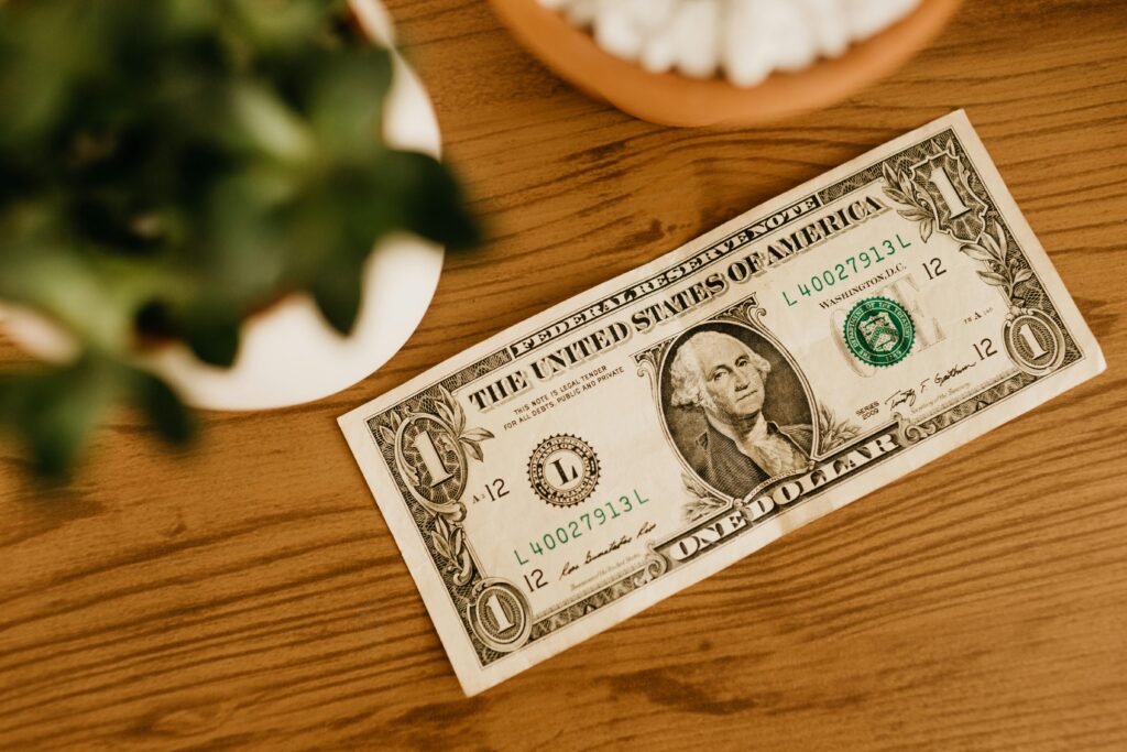 A US one dollar bill placed on a wooden table with a blurred plant nearby.